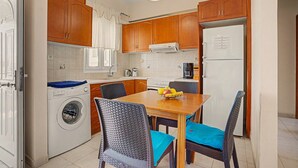 Een kleine keuken met houten kastjes, een wasmachine, een eettafel met stoelen en een koelkast.