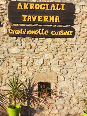 Un mur de pedra amb un senyal de fusta que diu 'AKROGIALI TAVERNA Cuina tradicional'.