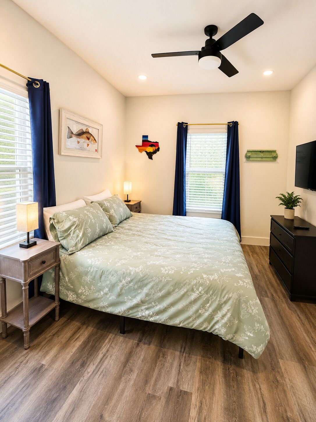 A bedroom with a bed, a ceiling fan, a nightstand with a lamp, a TV on a dresser, and a window with curtains.