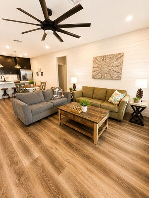 A modern living room with a ceiling fan, two sofas, a coffee table, and a kitchen in the background. - 3 BD Family Home | Extd Stays | Pets Ok (Aransas Pass)