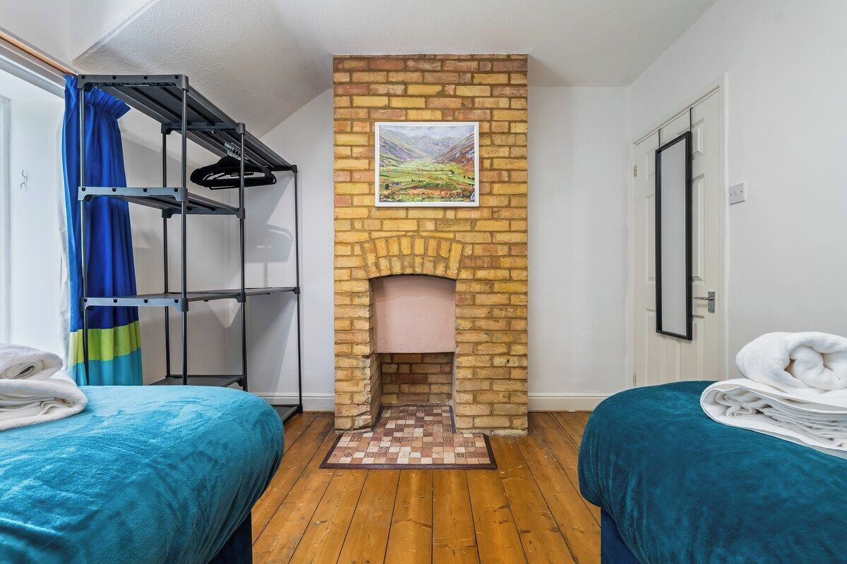 Een kamer met een open haard van baksteen, twee bedden met blauw beddengoed, een metalen plankenwand en een lijst met een landschapsafbeelding.