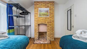 Een kamer met een open haard van baksteen, twee bedden met blauw beddengoed, een metalen plankenwand en een lijst met een landschapsafbeelding.