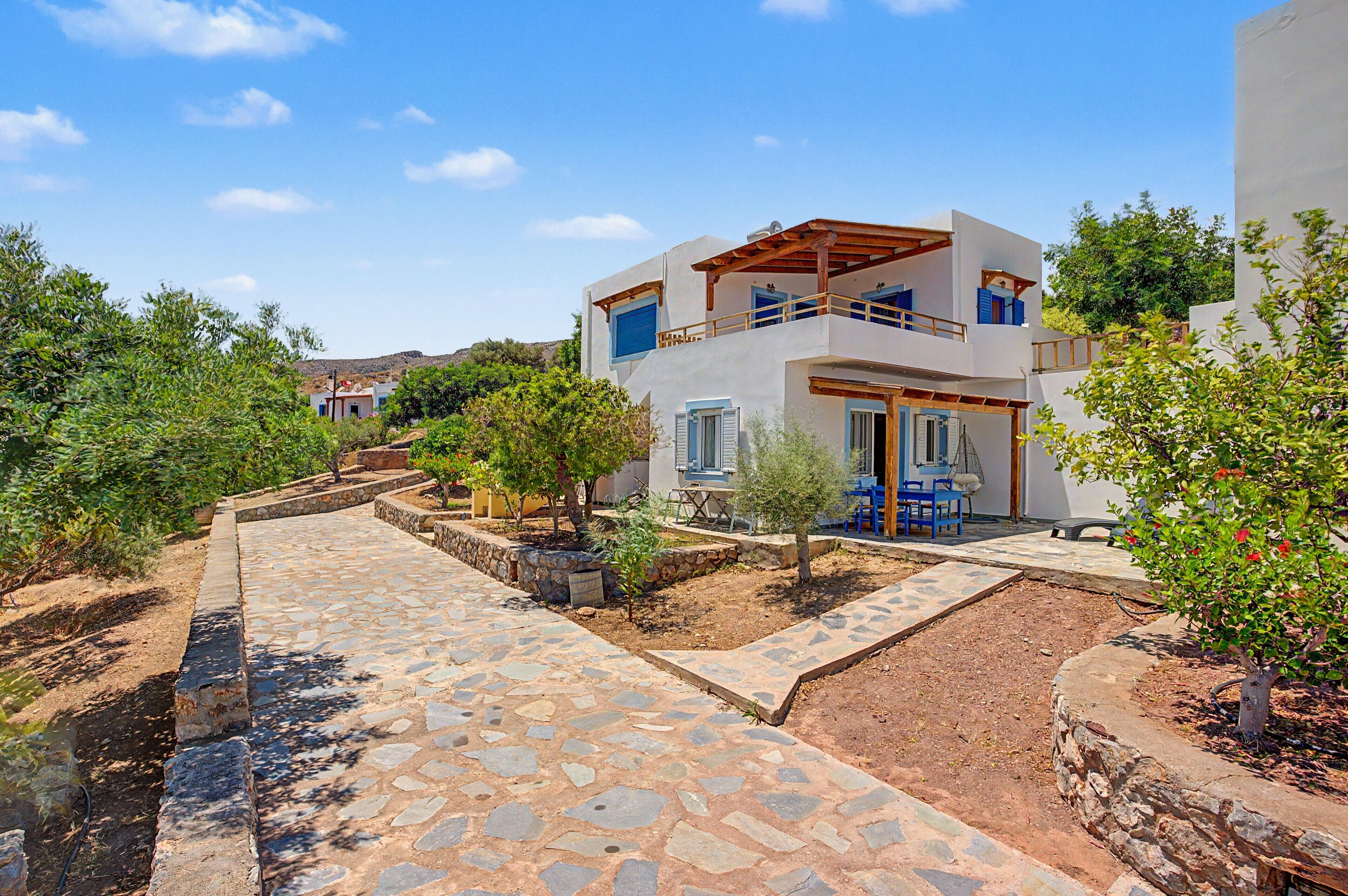 A two-story building with a stone-paved entrance, a balcony with blue shutters, and a garden area with trees and shrubs.