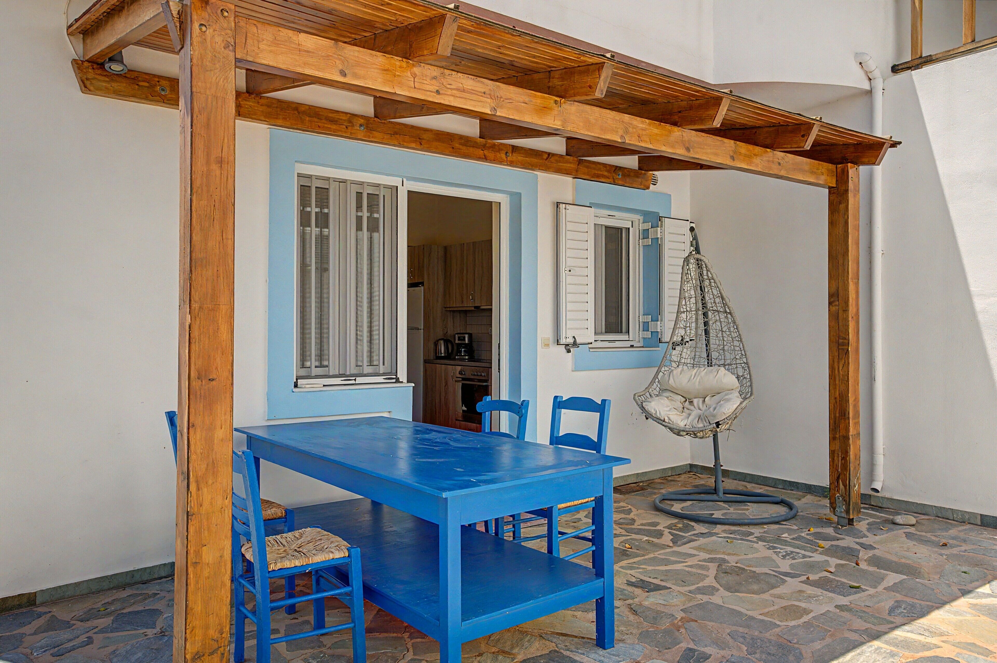 Une pièce avec une table et des chaises bleues, un fauteuil suspendu et une cuisine visible à travers une porte ouverte.