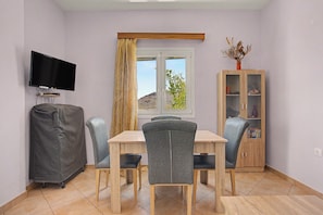 Ein Zimmer mit Esstisch, Stühlen, Fernseher, Schrank und einem Fenster mit Vorhängen.