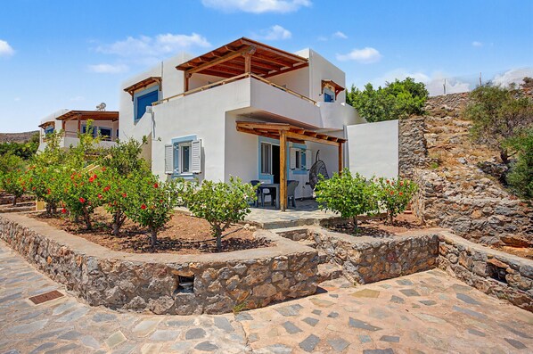 A modern two-story house with a stone foundation, wooden pergola, and a garden with flowering plants.