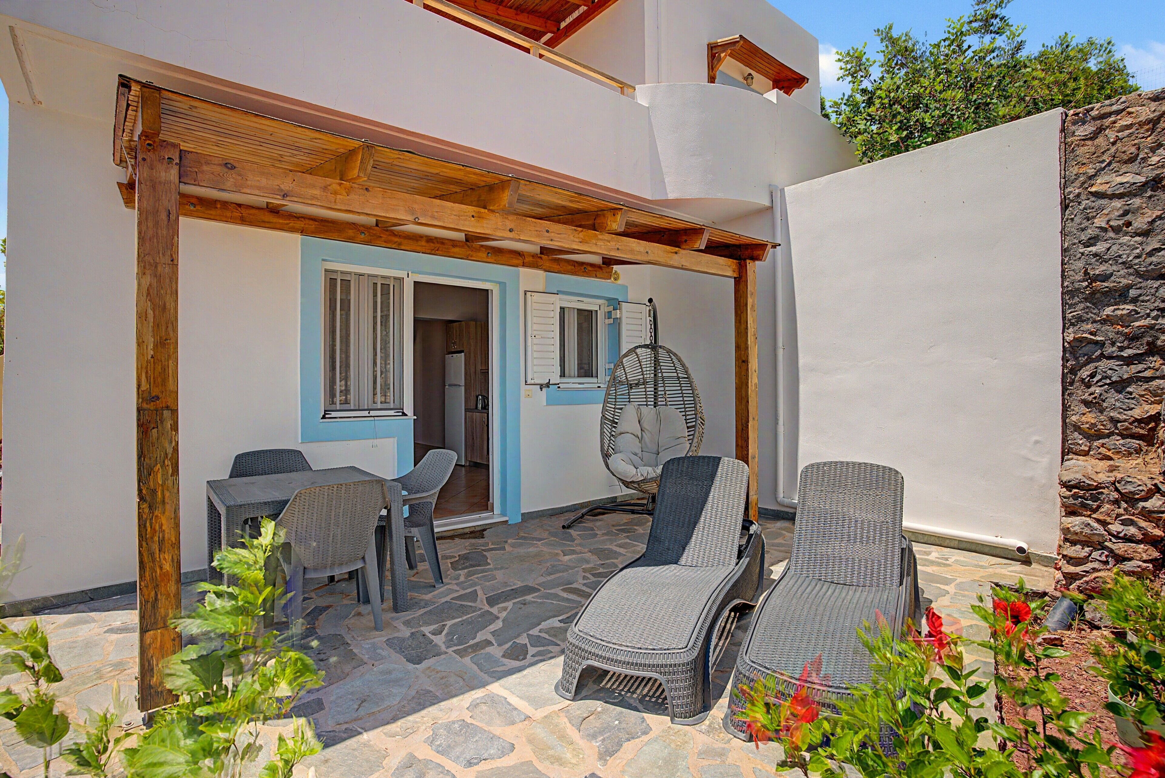 A covered outdoor seating area with wicker furniture, a stone floor, and a view of a kitchen through an open door.