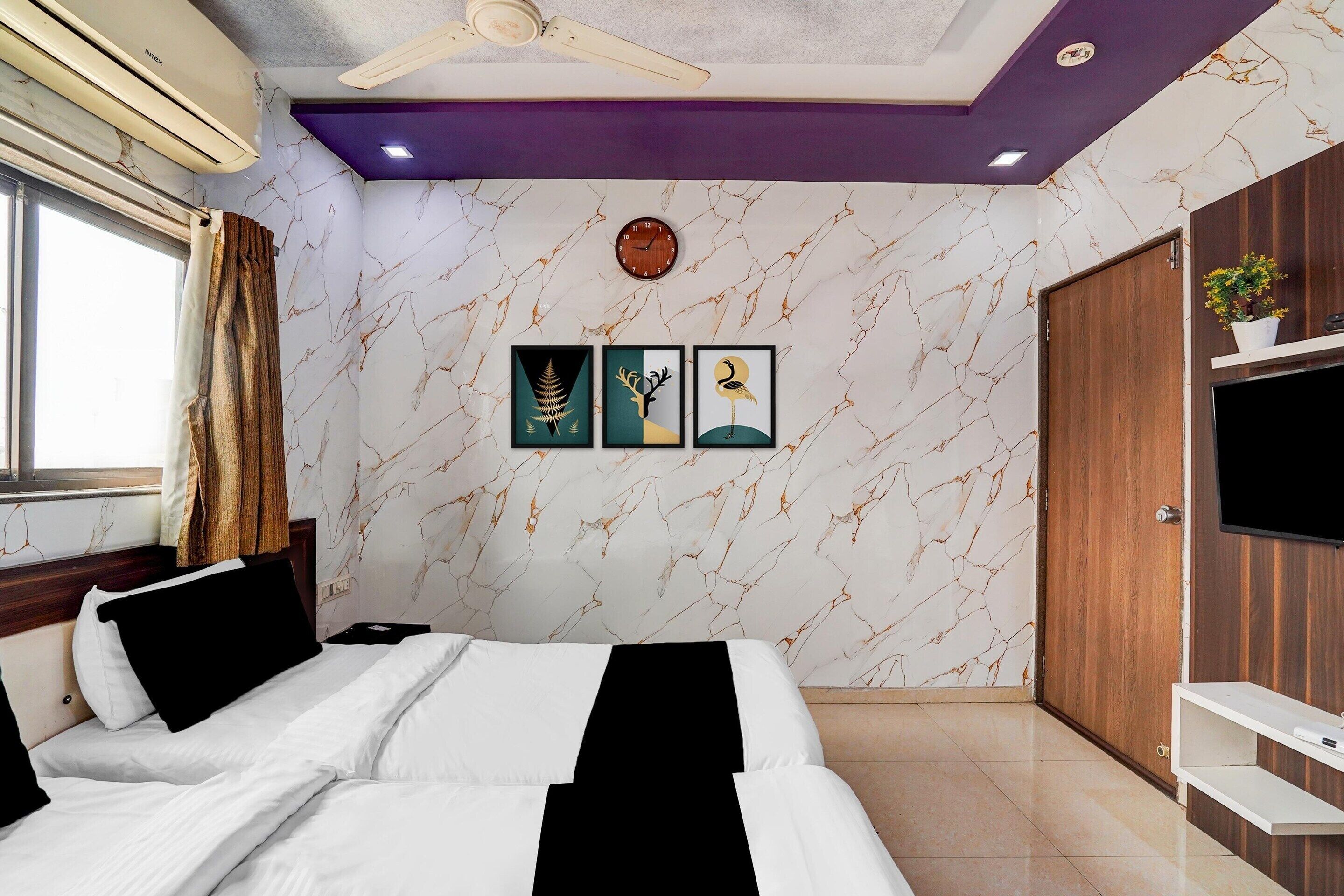 A hotel room with a bed, a ceiling fan, a clock, a TV, and a wooden door.