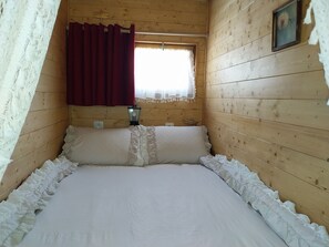 Habitación de cabaña de madera con una cama, una ventana con cortina roja, una lámpara de pared y un pequeño objeto decorativo.