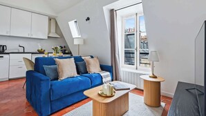 Ett modernt vardagsrum med en blå soffa, ett träbord och utsikt över byggnader genom ett fönster.