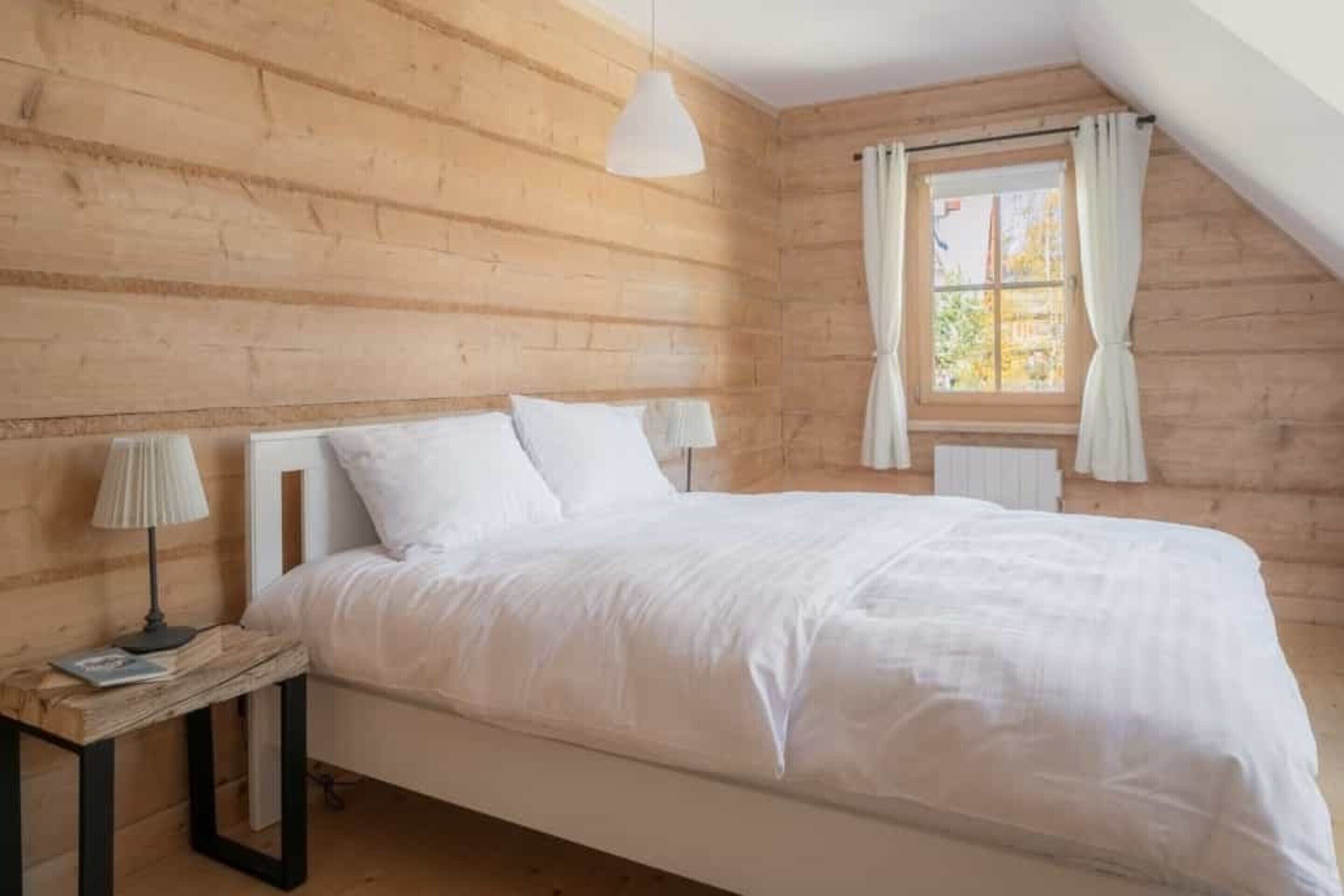 Une chambre avec un plafond en pente en bois, un lit avec une literie blanche, deux lampes de chevet et une fenêtre avec des rideaux.