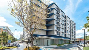A multi-story building with balconies, a curved facade, and a parking area with cars and a tree. - Modern Condo with Pool View D Condo Phuket C175 (ตำบล กะทู้)