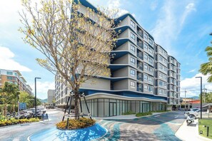 Un edificio de varios pisos con balcones, una fachada curva y un estacionamiento con coches y un árbol.