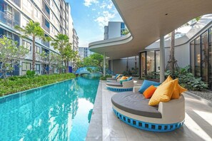 Zona de piscina con un diseño curvo de asientos, una piscina cristalina y edificios modernos al fondo.