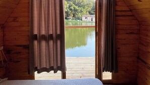 A room with a bed, wooden walls, a window, and a view of a lake. - Camping Mairiporã (Mairiporã)