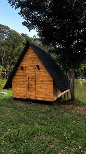 A wooden cabin with a pitched roof, situated on grass near a body of water, surrounded by trees. - Camping Mairiporã (Mairiporã)