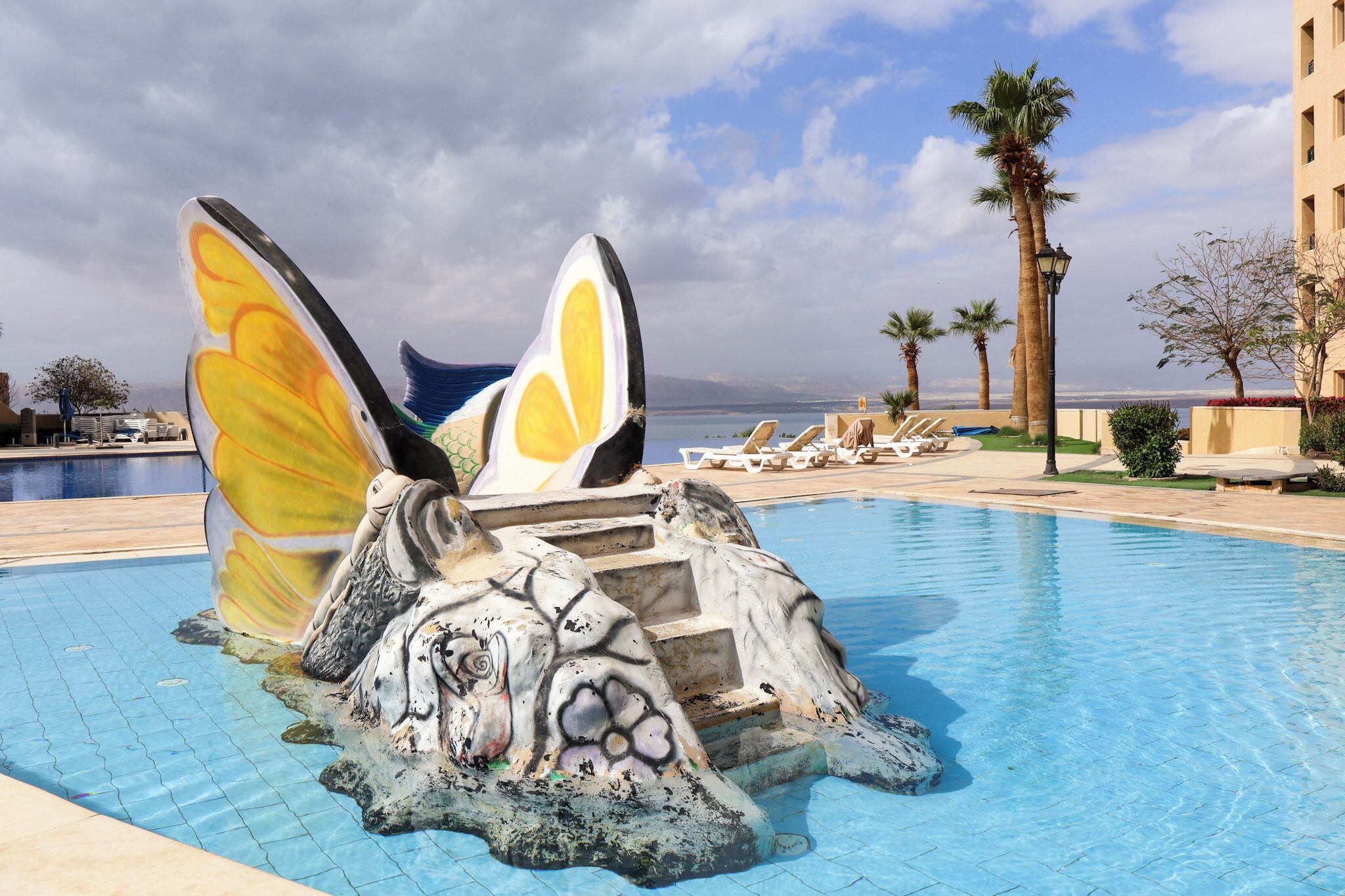 A pool with a decorative sculpture featuring wings and a face, surrounded by lounge chairs and palm trees.