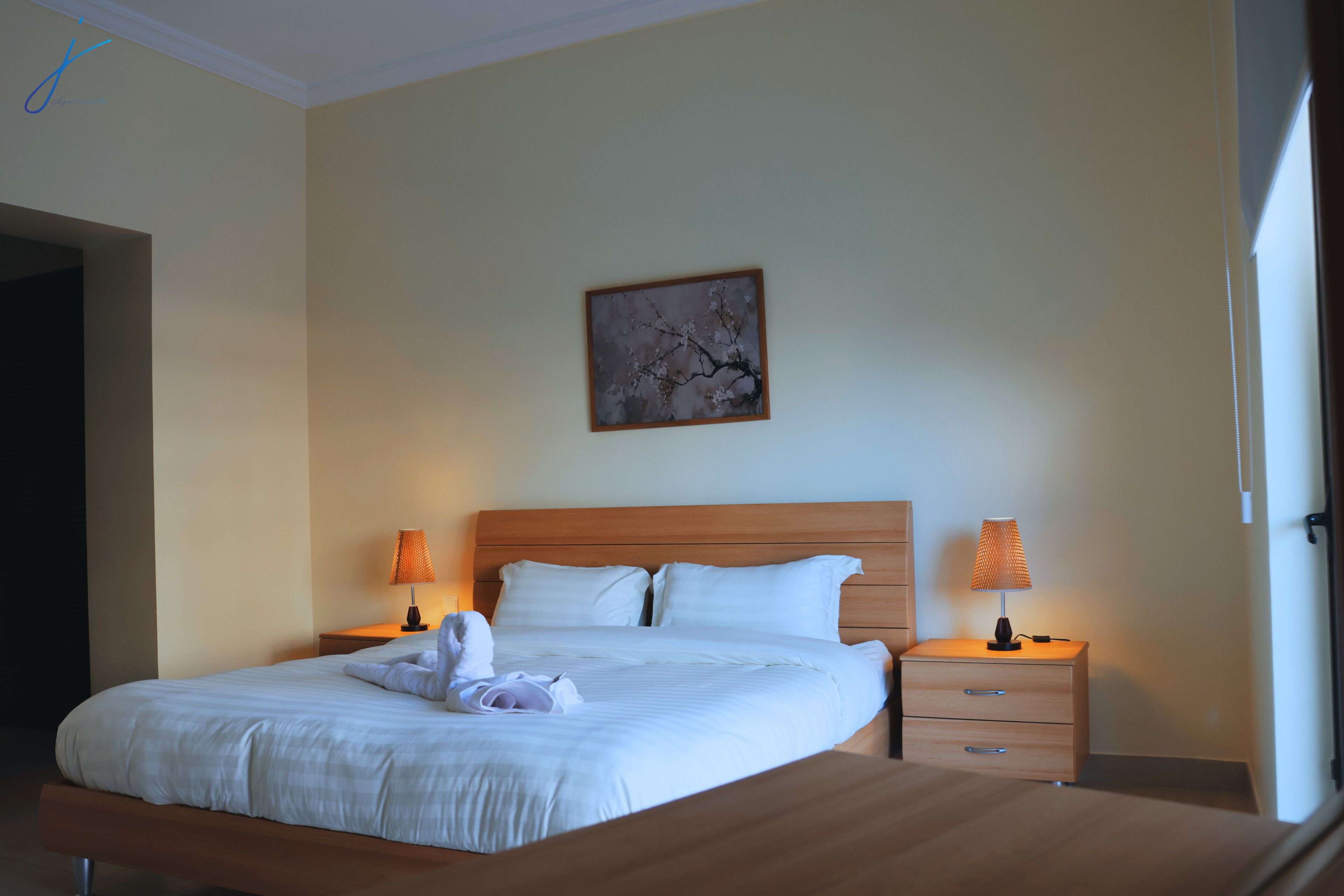 Une chambre d’hôtel comprenant un lit, deux tables de chevet avec des lampes, une tête de lit en bois, une œuvre d’art accrochée au mur et une porte.