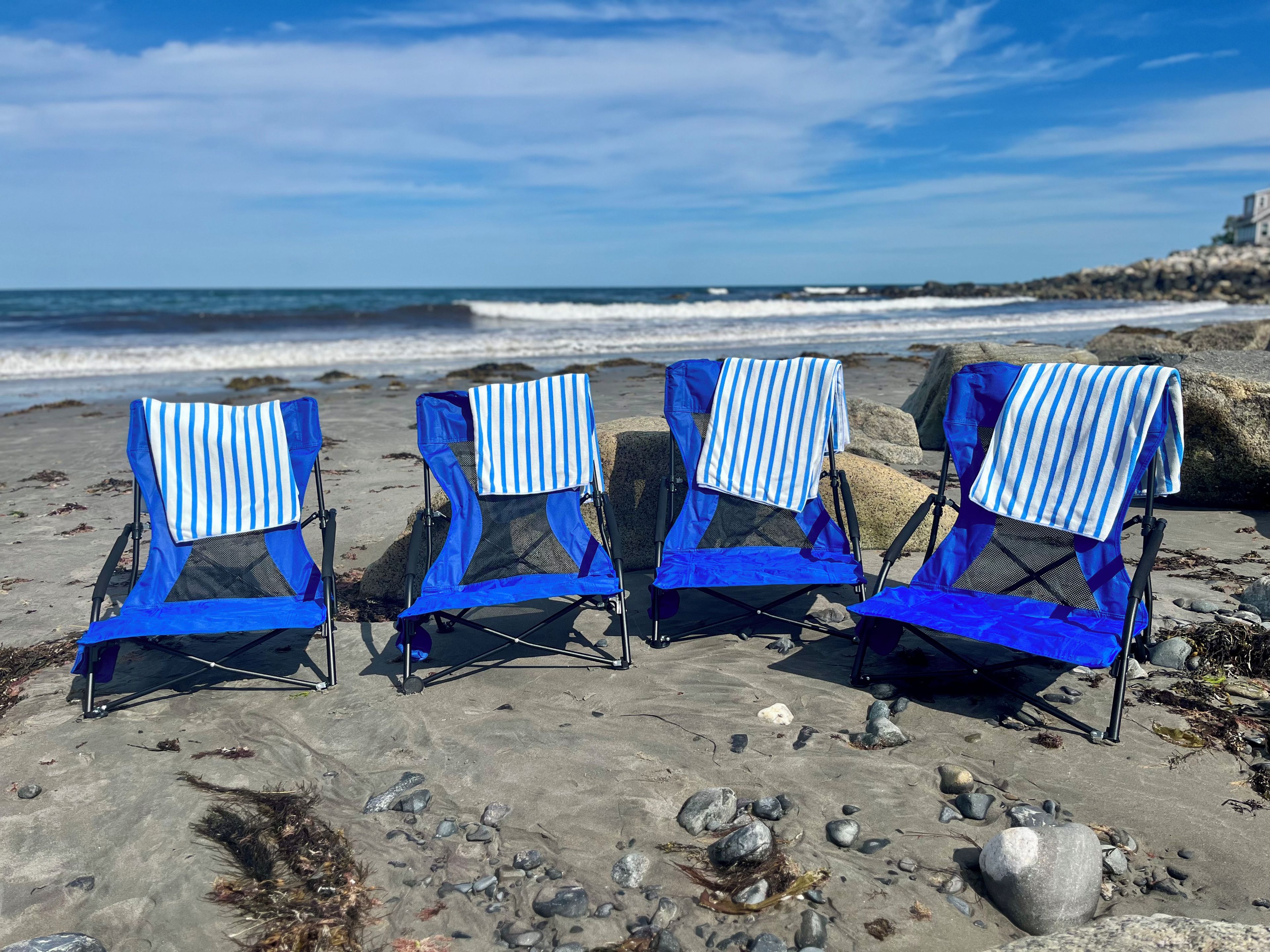 Four beach chairs with blue and white striped towels, arranged on a rocky beach with the ocean in the background.