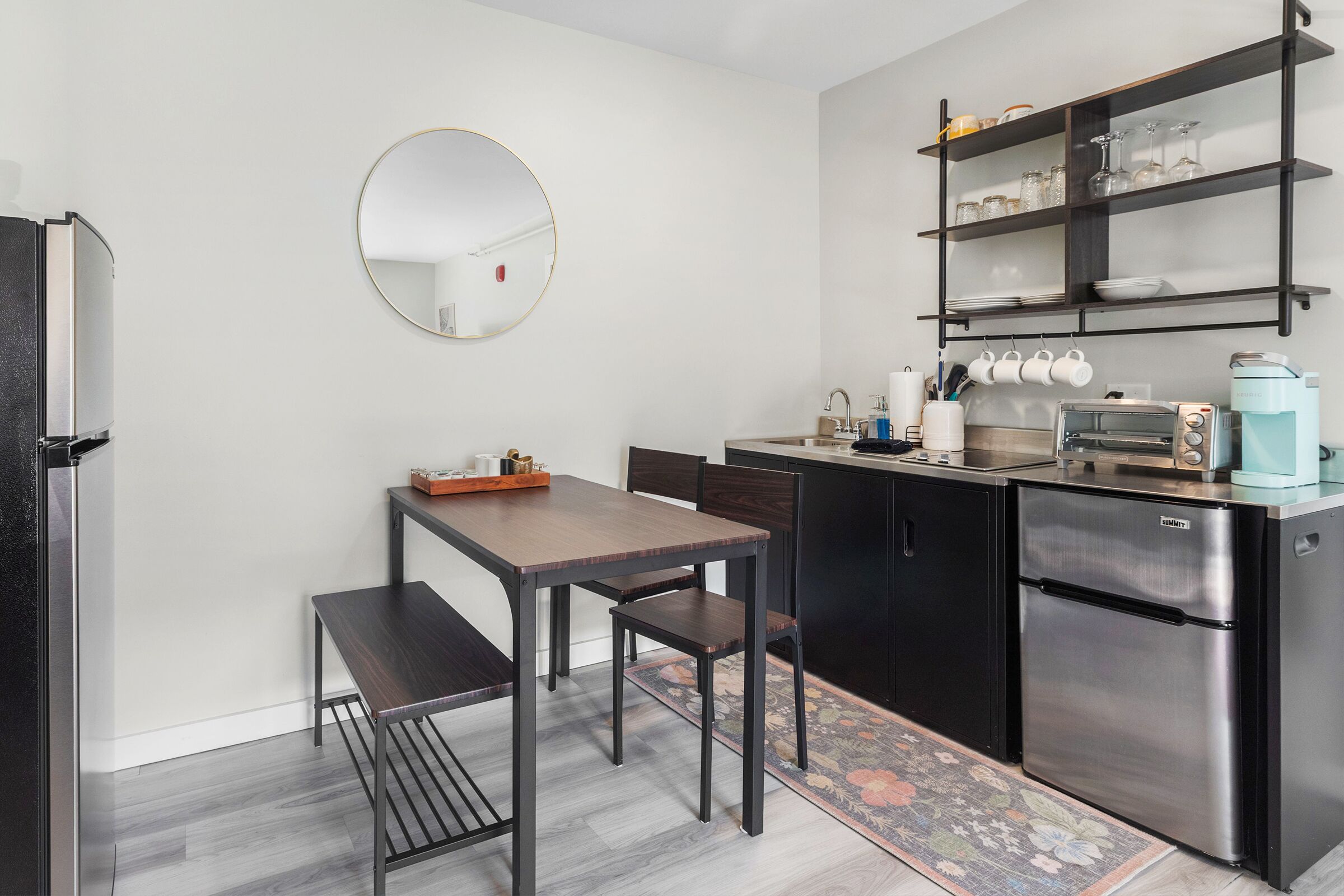 A compact kitchen with a round mirror, a dining table with chairs, a coffee machine, and a refrigerator.