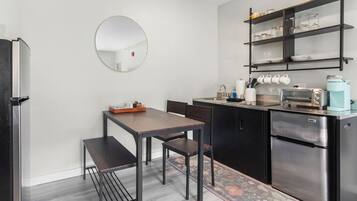 A compact kitchen with a round mirror, a dining table with chairs, a coffee machine, and a refrigerator.