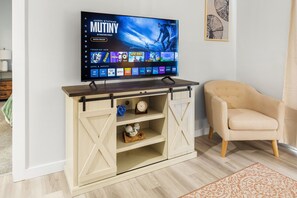 A flat-screen TV mounted on a wooden entertainment center with a sliding barn door, a beige armchair, and a patterned rug on a wooden floor. - Ocean Mist Condo- Sleeps 4-Beach View- Walk to Town-Parking- Deck (Hampton)