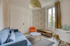 Ein modernes Wohnzimmer mit einem blauen Sofa, einem orangefarbenen Sessel, einem kleinen weißen Tisch und einer Wand mit Marmoroptik.