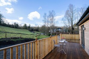 Eine Holzterrasse mit Tisch und Stühlen, Blick auf ein grünes Feld, und ein Gebäude im Hintergrund.