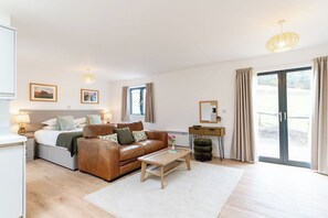 Ein modernes Hotelzimmer mit einem Bett, einer Ledersofa, einem Holztisch, einem kleinen Beistelltisch, einem Spiegel und einer Schiebetür zum Außenbereich.