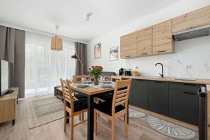 A modern kitchen with wooden cabinets, a dining area with a table set for two, and a large window with sheer curtains. - Cozy Apartment Near the Beach | Parking | Terrace (Międzywodzie)