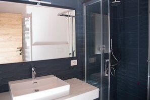 Ein modernes Badezimmer mit Duschkabine aus Glas, einem weißen Waschbecken und einer Holzvertäfelung über dem Spiegel.