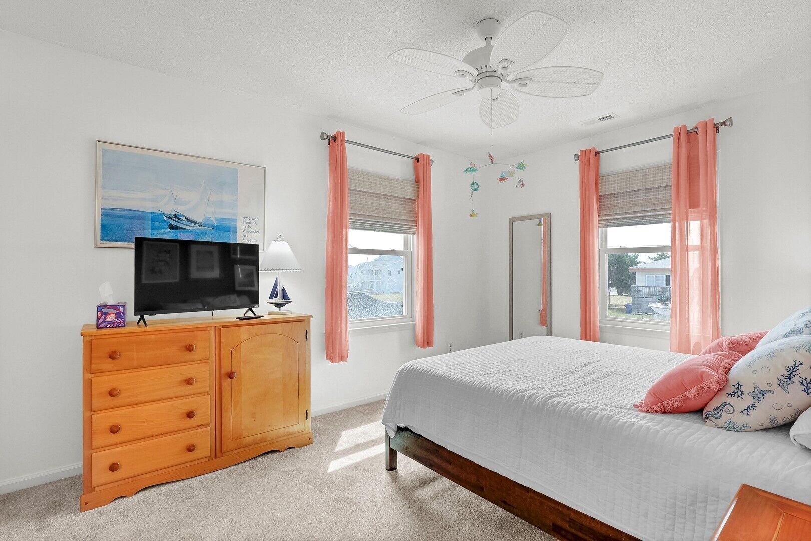 A bedroom with a bed, a TV on a wooden dresser, a ceiling fan, and pink curtains.
