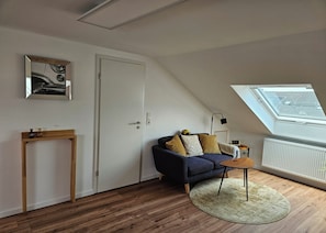 A room with a loft-style ceiling, a sofa with cushions, a small table, a lamp, and a radiator. - Quiet attic studio in Cologne-Holweide, a 2-minute walk to the S-Bahn station for Deutz/Messe (Köln)