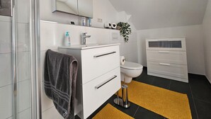 A modern bathroom with a glass shower enclosure, a white sink with a mirror above, a toilet, and a white cabinet with drawers. - Quiet attic studio in Cologne-Holweide, a 2-minute walk to the S-Bahn station for Deutz/Messe (Köln)
