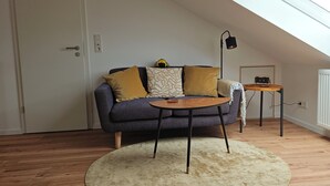 A loft-style room with a sloped ceiling, a dark grey sofa with patterned cushions, a small wooden side table, and a round rug. - Quiet attic studio in Cologne-Holweide, a 2-minute walk to the S-Bahn station for Deutz/Messe (Köln)