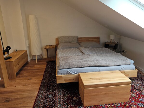 A bedroom with a sloped ceiling, a bed with striped bedding, a wooden nightstand, a bedside table, a lamp, a small wooden dresser, and a patterned rug.