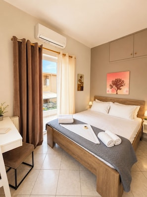 A hotel room with a bed, a desk, a chair, a window with curtains, an air conditioning unit, and a painting on the wall. - Villa Morea With Pool Near Chania City (Chania)