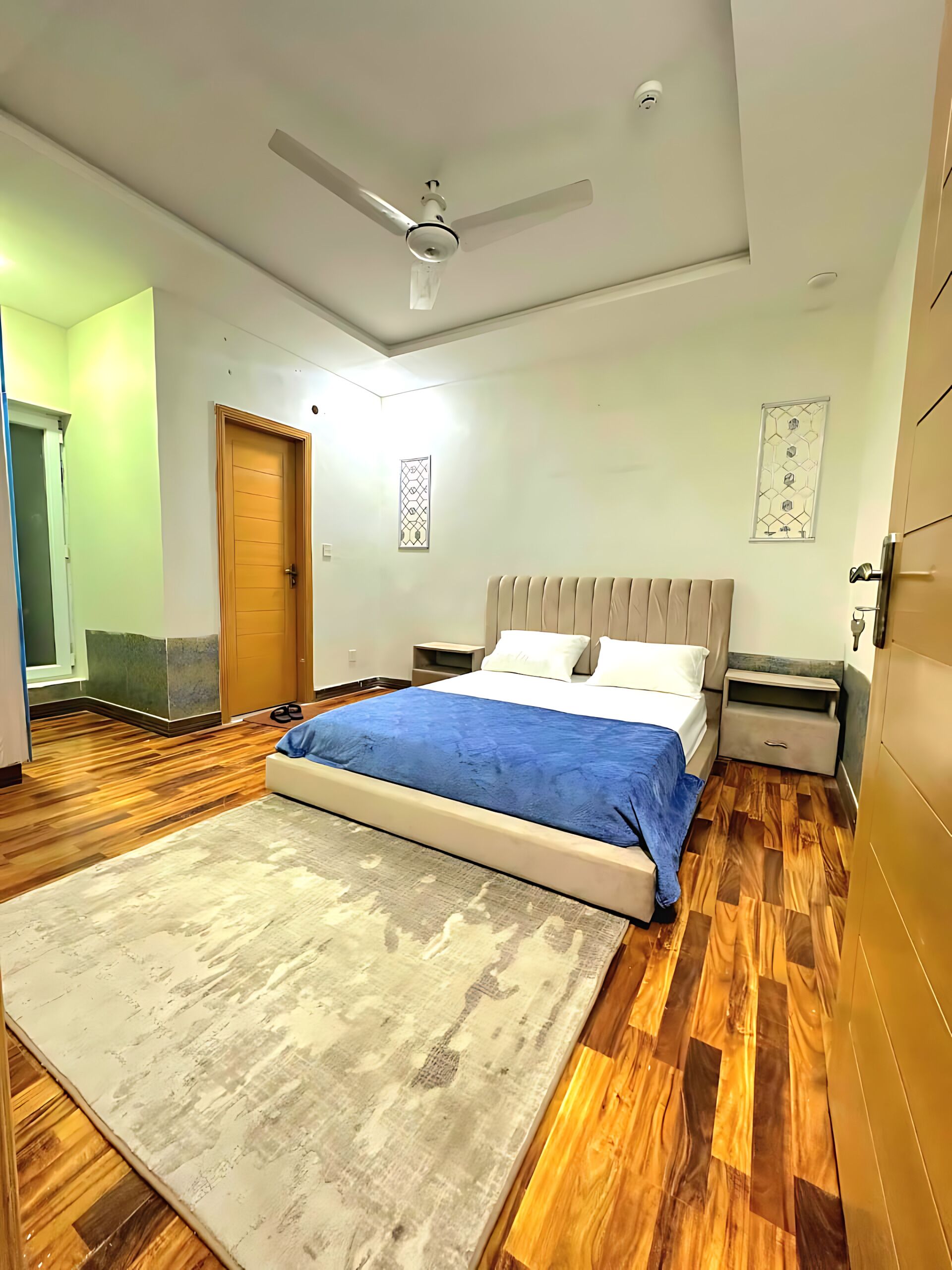 Quarto com cama, mesa de cabeceira, ventilador de teto e piso de madeira.
