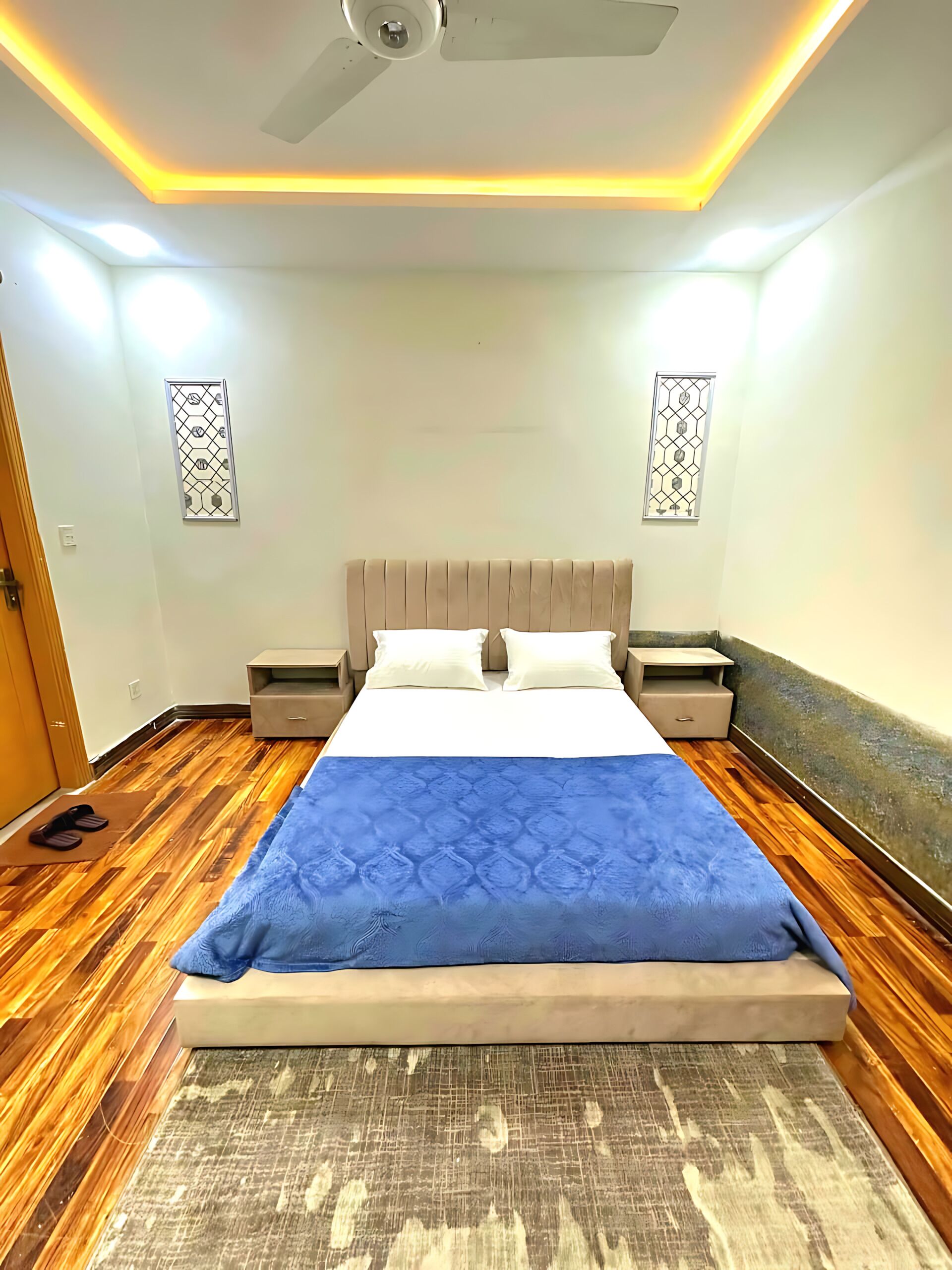 A bedroom with a bed, nightstands, a ceiling fan, and two wall-mounted windows.