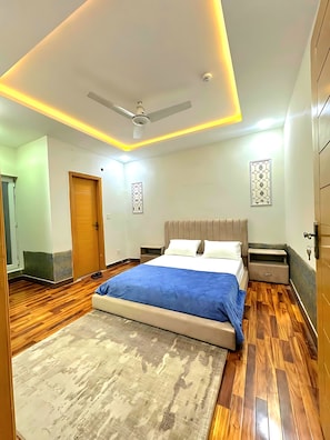 Une chambre à coucher comprenant un lit, un ventilateur de plafond, une table de chevet, une armoire et une porte.