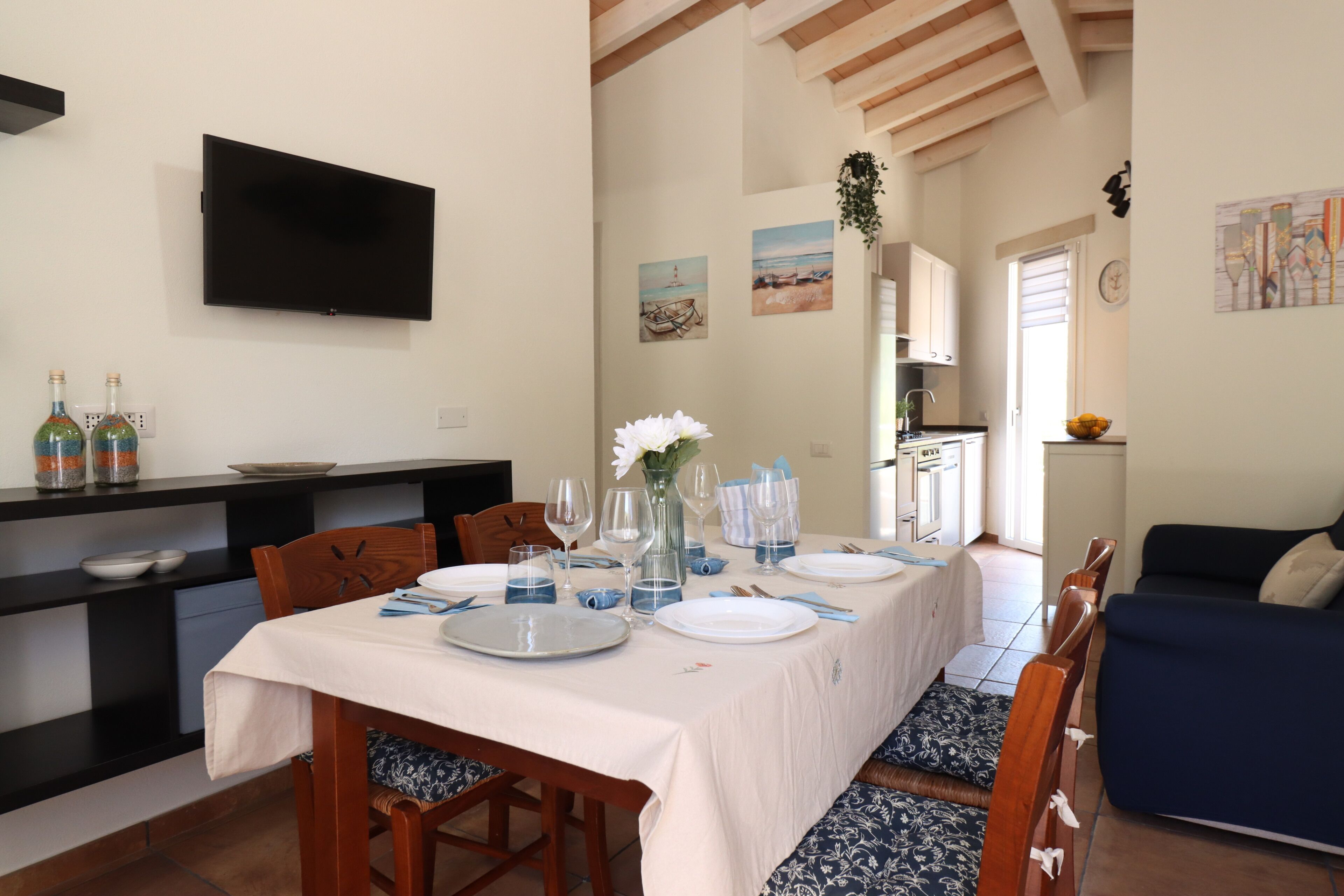 A dining area with a table set for four, a TV mounted on the wall, and a kitchen in the background.