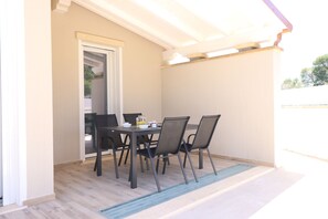 A covered patio with a table and chairs, a door leading to an interior space, and a wooden floor. - Mar | House in Solanas between sea and nature (Cuili Murvoni)