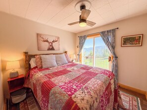 Een slaapkamer met een groot bed, een plafondventilator, een raam met gordijnen, een schilderij aan de muur en een nachtkastje met een lamp.