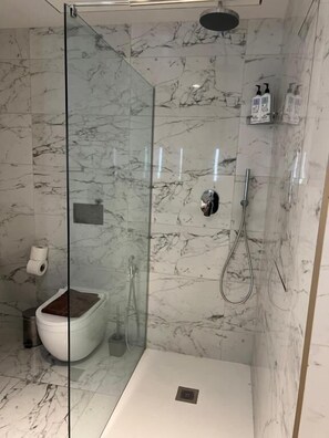 A glass-enclosed shower with a marble wall, a toilet, and a small shelf with toiletries. - Mercury tower suites by Asignatures (St. Julian's)