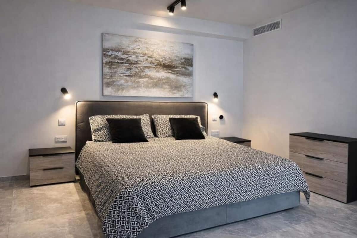 Une chambre moderne avec un grand lit, deux tables de chevet, une commode, une coiffeuse et un luminaire fixé au mur.