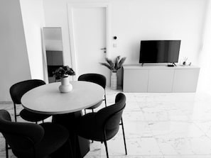 A modern dining area with a round table, six chairs, a vase with dried flowers, a flat-screen TV on a white cabinet, and a potted plant.