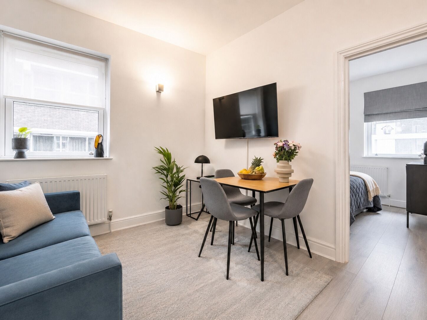 Ett modernt vardagsrum med en blå soffa, ett matbord med stolar, en krukväxt och en platt-TV som är monterad på väggen.