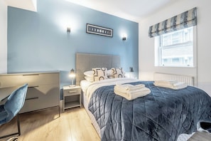 A bedroom with a bed, bedside table, lamp, and a window with curtains. - Kula London Tower Bridge (London)