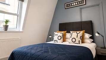 Un dormitorio con cama, almohadas, una mesita de noche, lámpara, ventana con planta y un cuadro colgado en la pared.