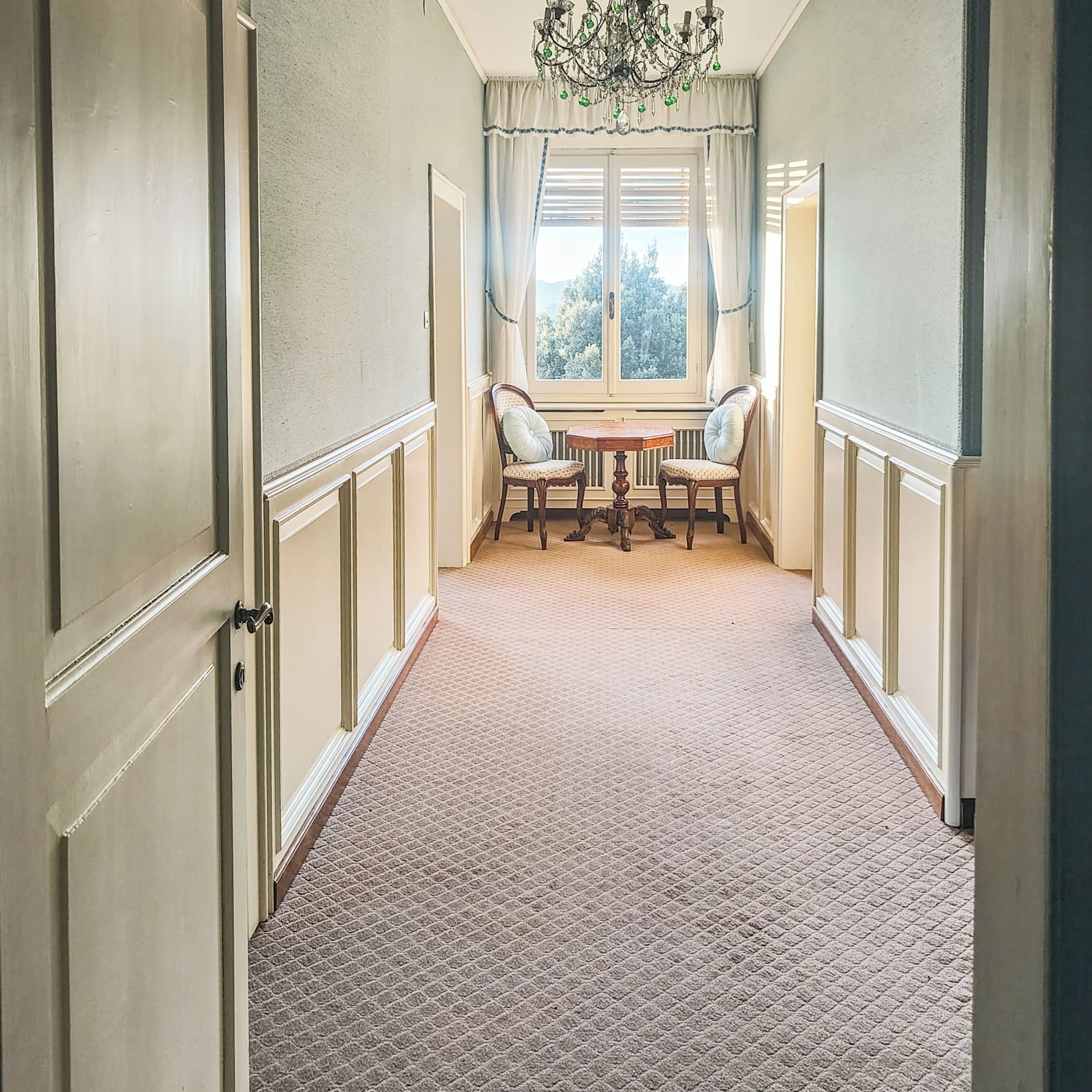 A hallway with a carpeted floor, a wooden table with chairs, a window with curtains, and a chandelier.